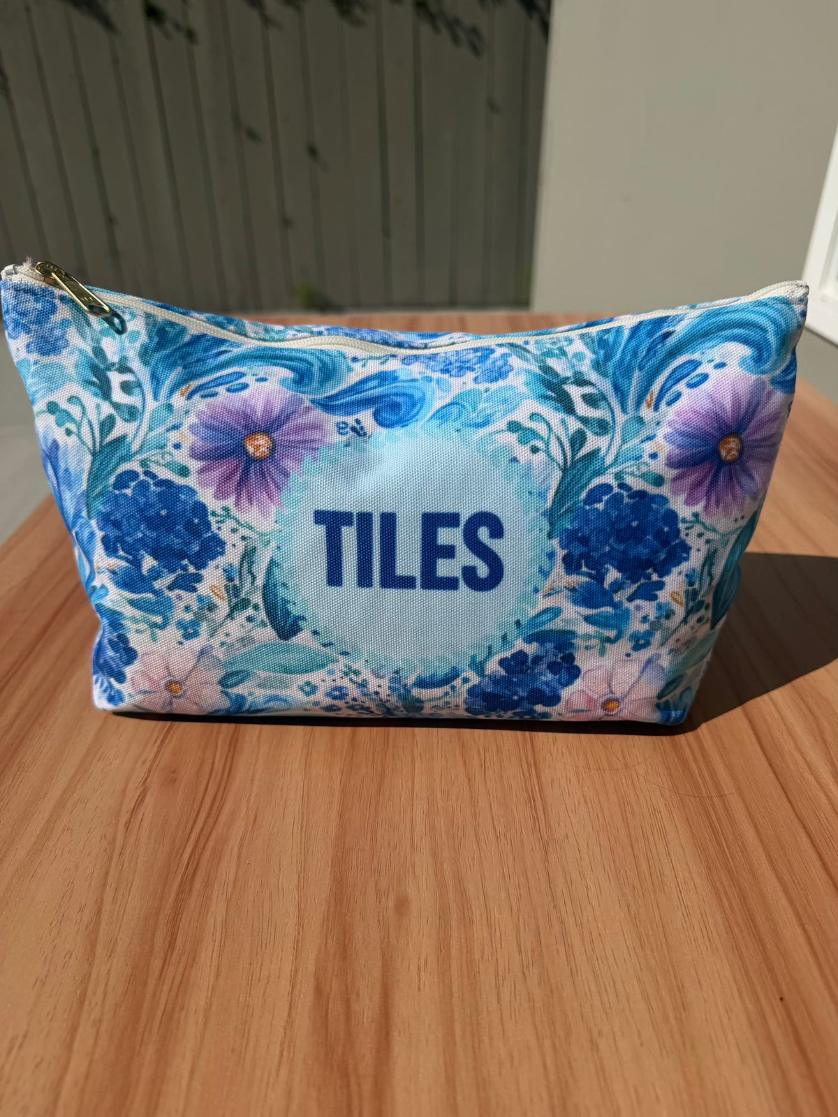 Blue and purple floral zippered pouch printed with TILES in navy lettering, standing on a wooden table