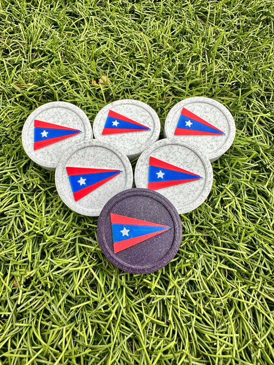 Six round backgammon checkers rest on grass, mostly white marble with one deep purple piece, each stamped with a custom logo.