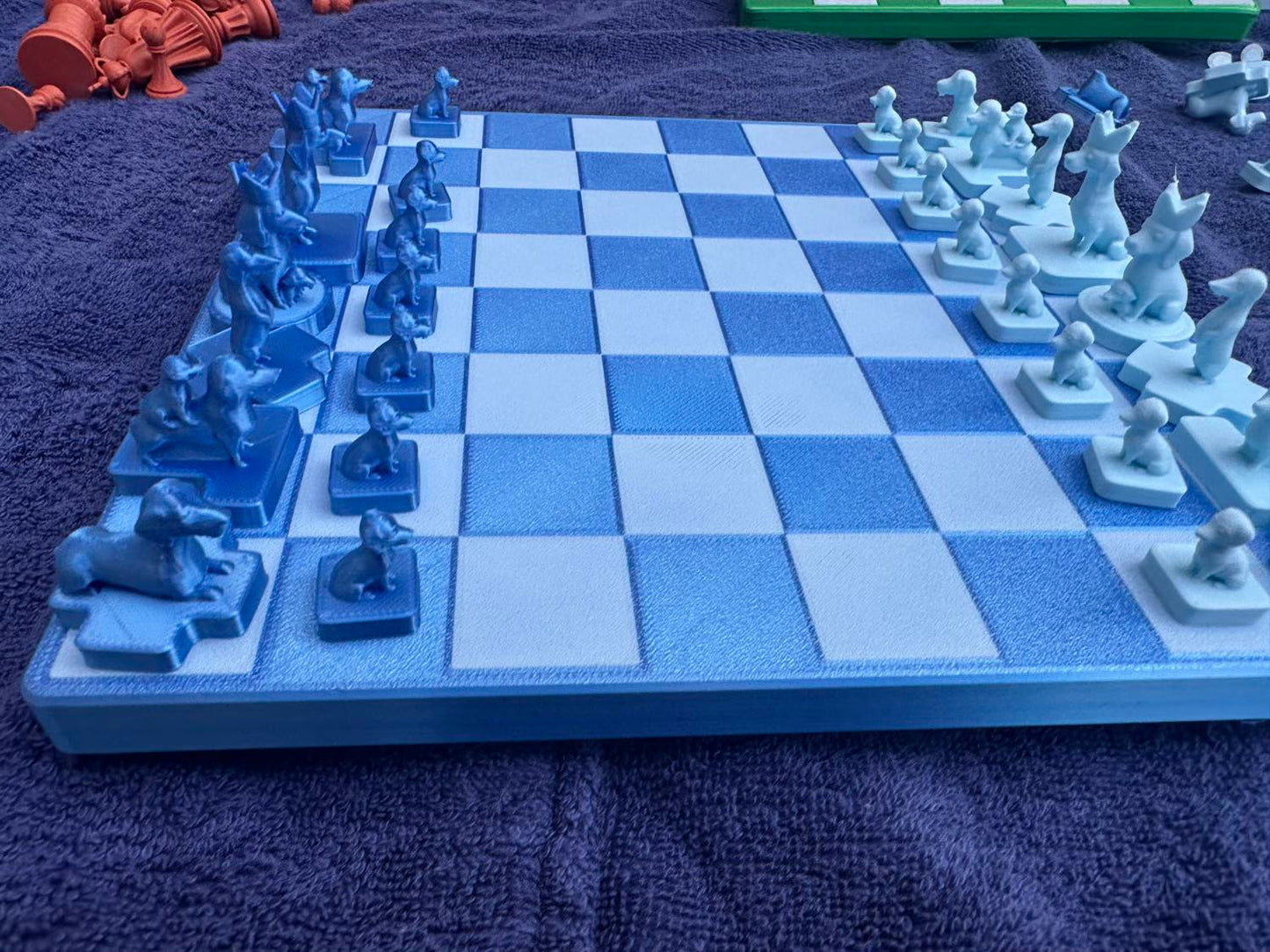 A blue Dachshund Travel Chess board holds its dog-shaped pieces mid-setup, orange pieces and a green rack blurred behind.