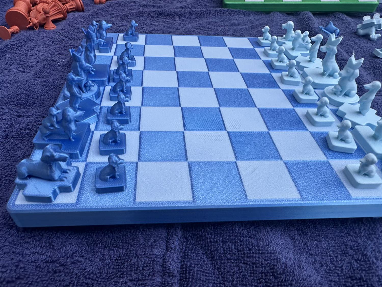 A blue Dachshund Travel Chess board holds its dog-shaped pieces mid-setup, orange pieces and a green rack blurred behind.