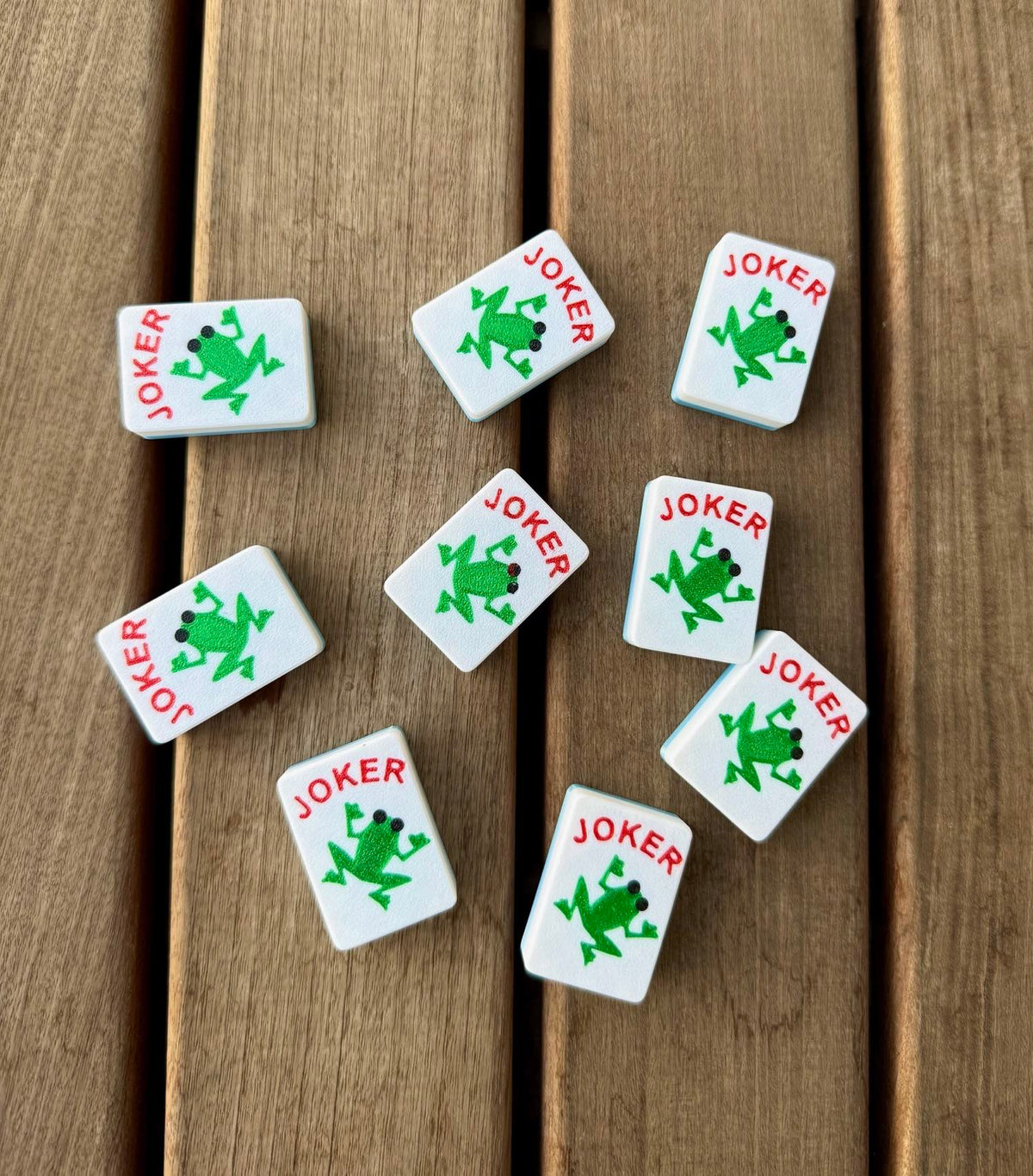 Eight Custom Jokers - Frogs tiles, each stamped with a green frog, scatter across a wood deck.
