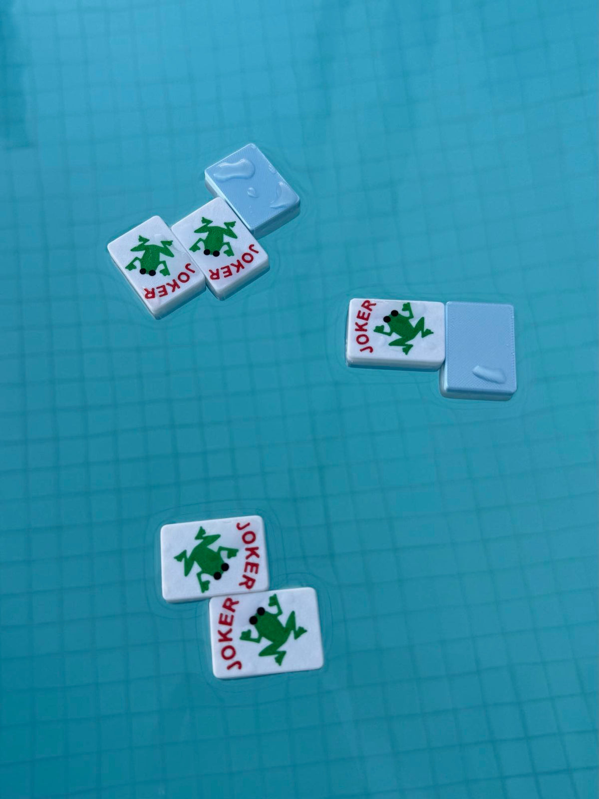Three green frog Joker mahjong tiles floating on the surface of a blue tiled pool