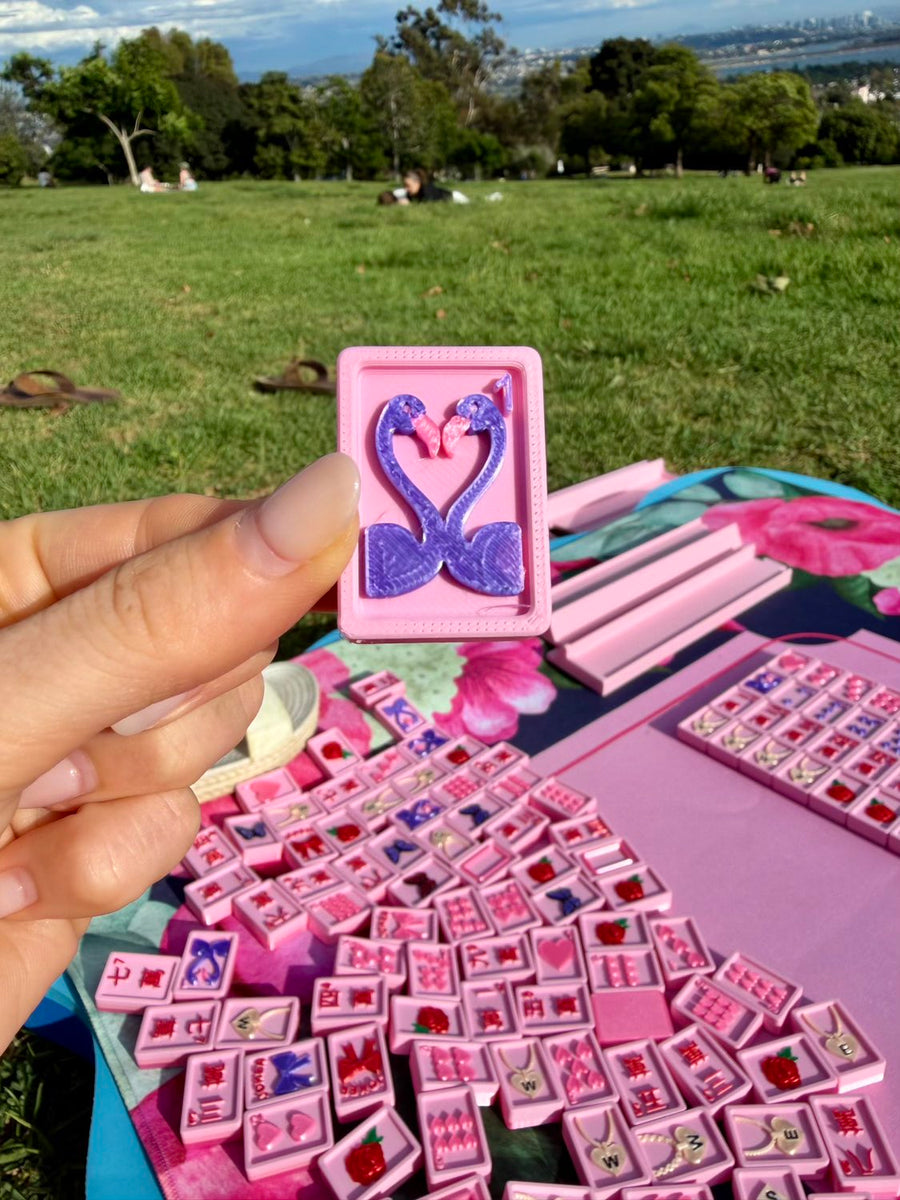 A hand holds the Flamingo joker tile, two flamingos forming a heart, over a blanket of pink tiles