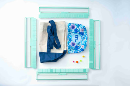 A styled flat-lay of a canvas tote bag with navy blue handles and trim, a blue floral zippered pouch with 'TILES' printed on it, and several mint green plastic grid tools arranged around it on a white background.