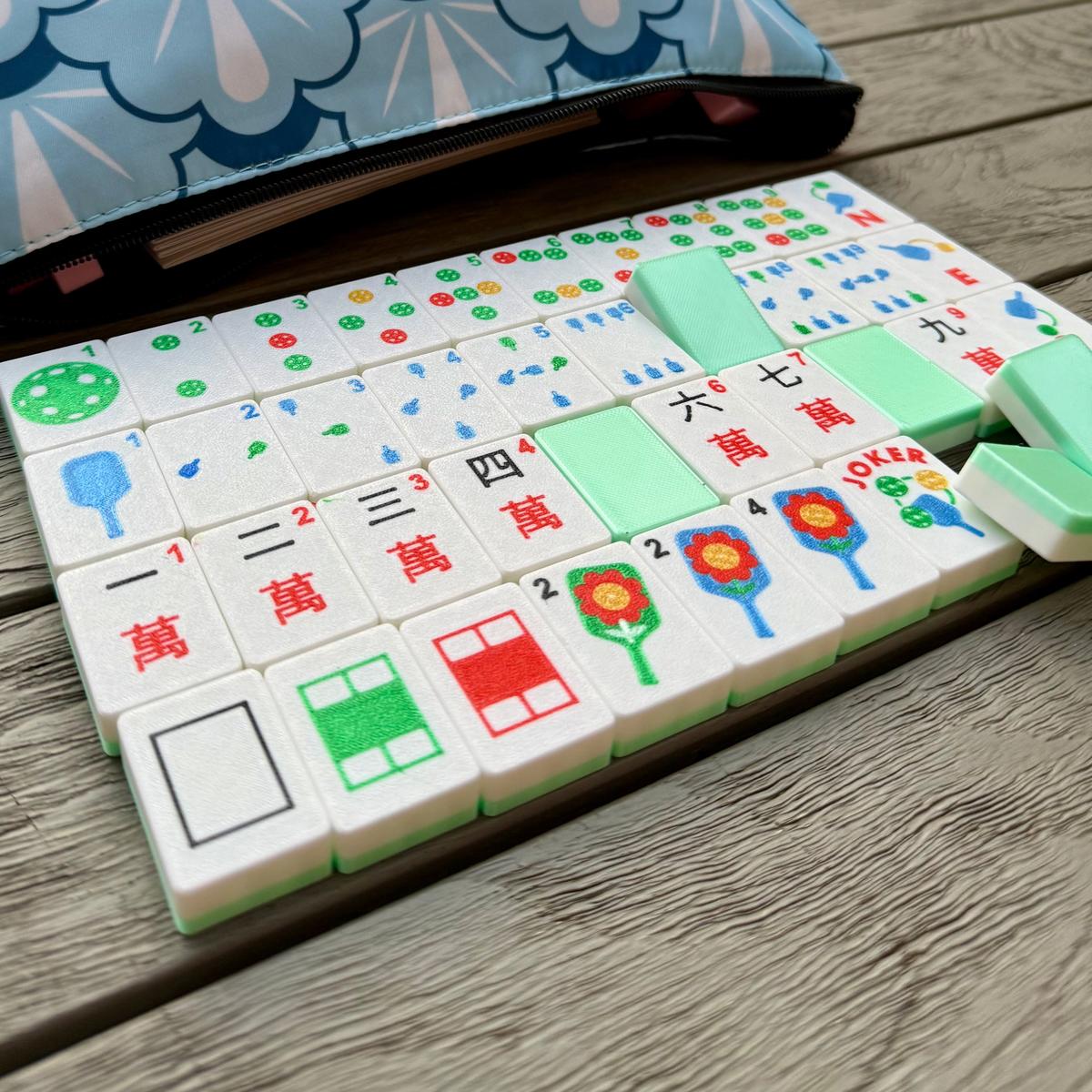 Pickleball Mahjong set with green tiles neatly arranged beside a blue carrying case, colorful American Mahjong game pieces by Spools

