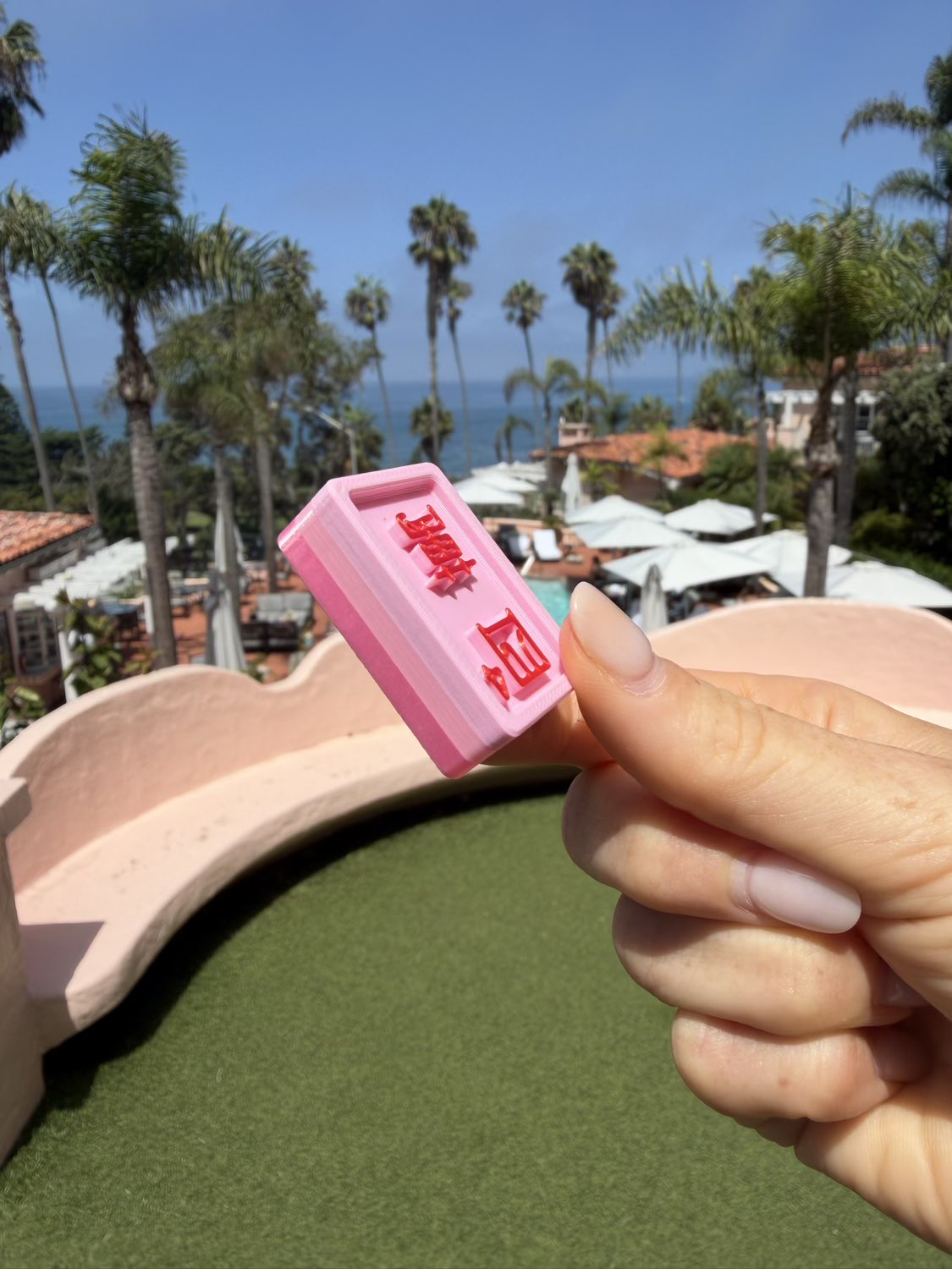 A hand holding a small pink rectangular Mahjong Tile with red characters, photographed outdoors with a beach and palm trees in the background.