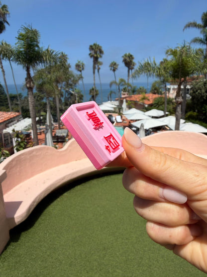 A hand holding a small pink rectangular Mahjong Tile with red characters, photographed outdoors with a beach and palm trees in the background.