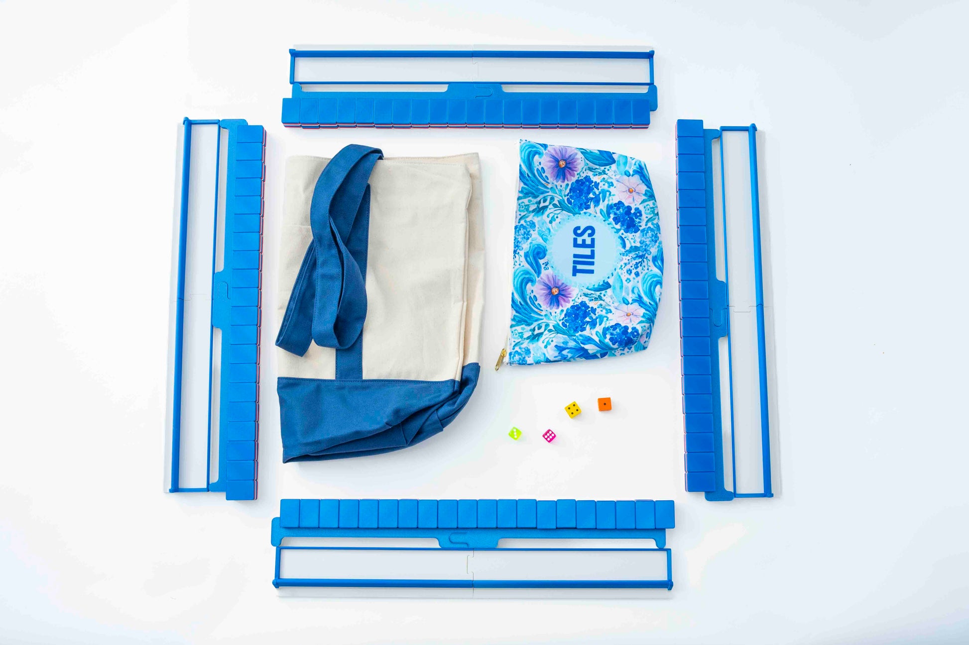 A flat-lay product shot of a personalized tote bag set on a white background, featuring a beige canvas tote with a blue fabric shoulder strap, a blue floral drawstring pouch printed with the name 'TILE', and multiple blue plastic organizer clips or dividers arranged around it.
