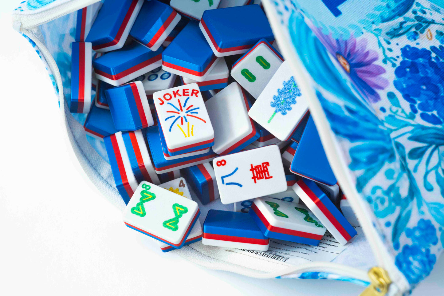 A close-up of Mahjong tiles spilling out of a blue and white floral-patterned zippered pouch on a white background.