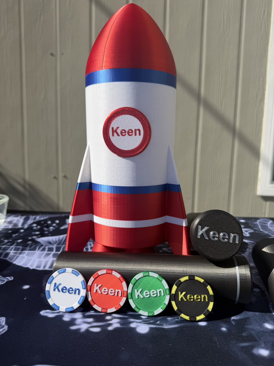 A red, white, and blue rocket-shaped mug or cup with the word 'Keen' printed on the front, displayed upright on a dark patterned tablecloth with matching round tokens in front.