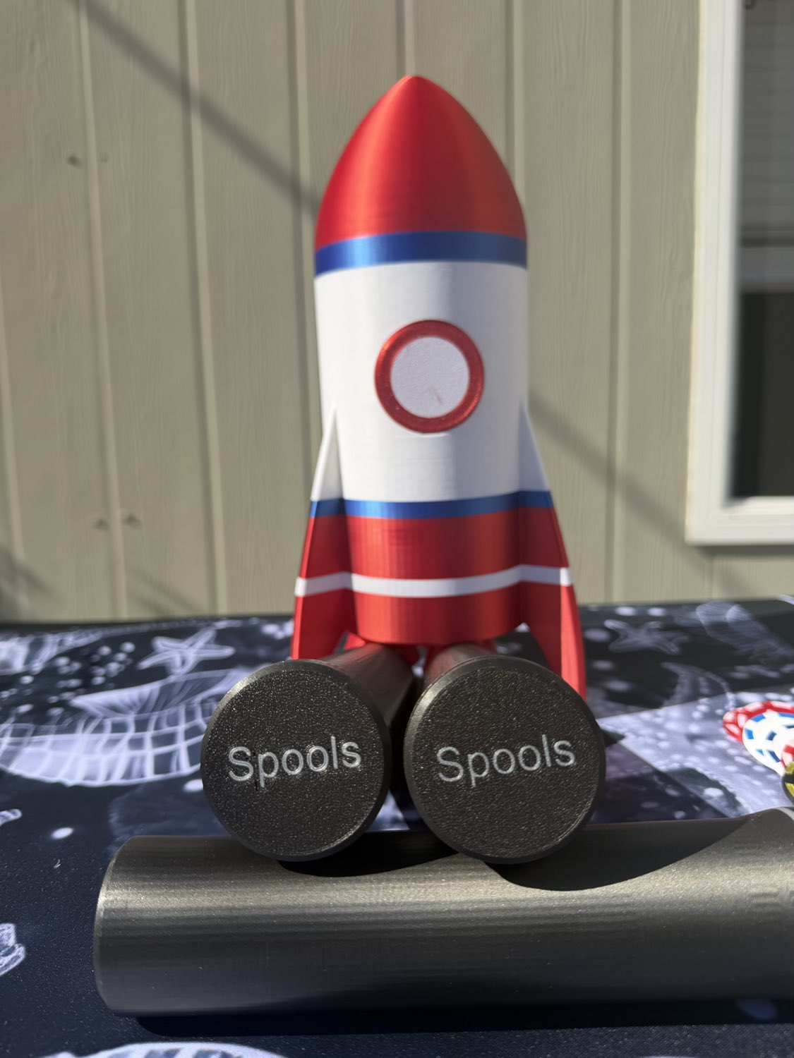 A decorative rocket-shaped product with a red, white, and blue design, standing on two dark round weights labeled 'Spools' on a metallic bar. The background shows a beige paneled wall and a window.
