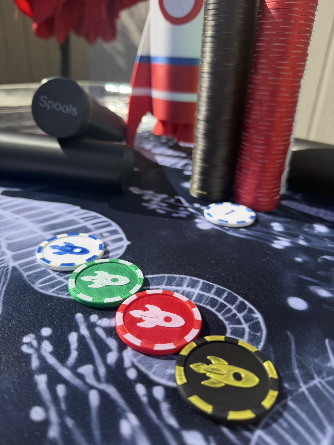 A close-up of a tabletop gaming setup with several round poker-style chips in red, green, blue, black, and yellow arranged on a dark printed mat. A black cylindrical object labeled 'Spools' is visible in the background, along with stacks of red, black, and white chips and a rocket-shaped item.