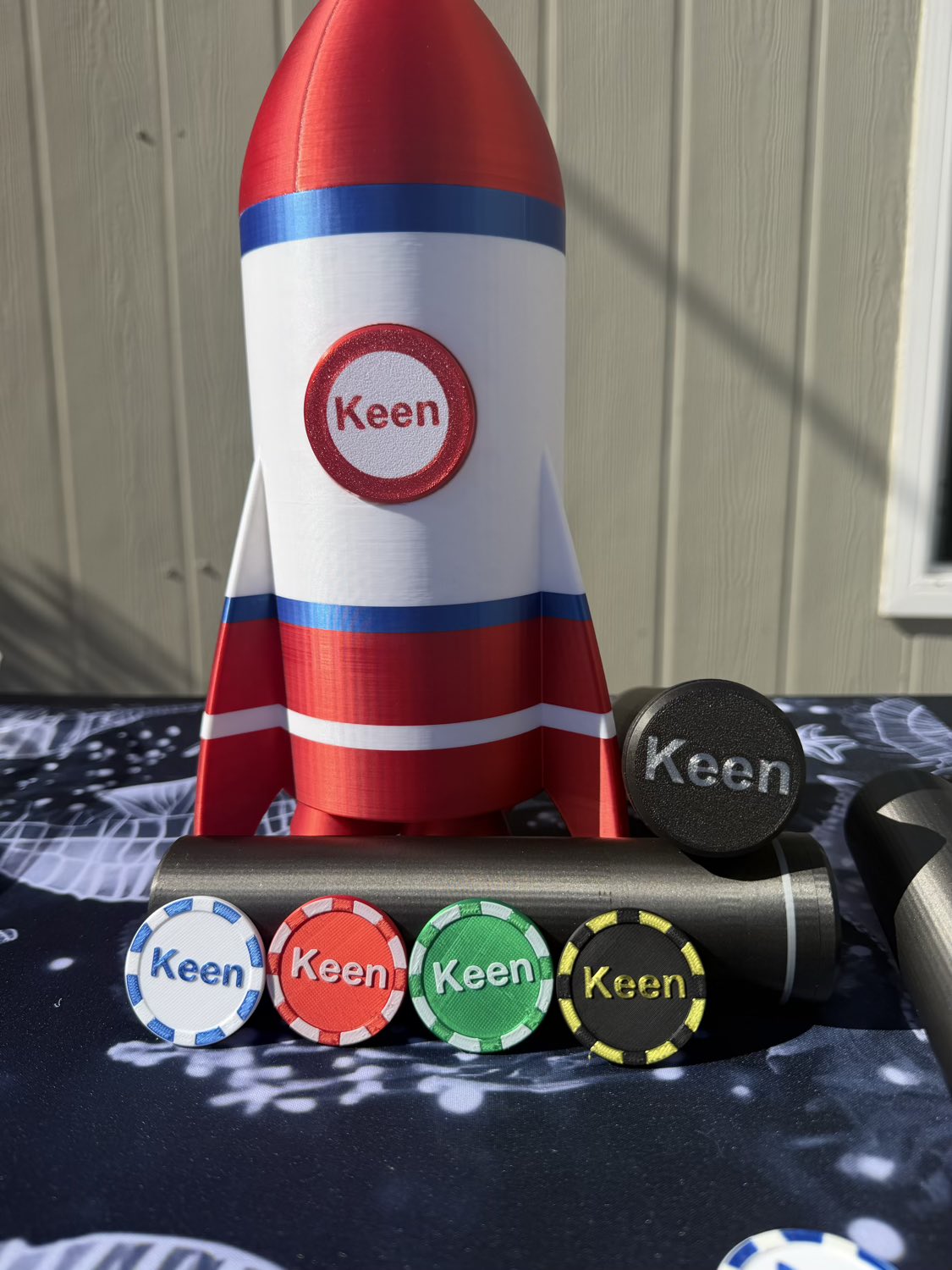 Personalized rocket-shaped game pieces with the name 'Keen' printed on them, displayed on a dark patterned surface in front of a light wood-paneled wall.