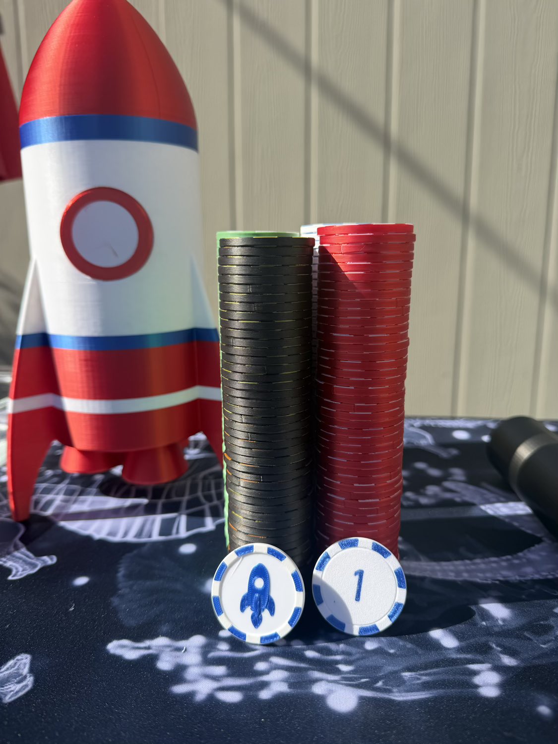 A set of gaming chips and a rocket theme prop on a patterned tablecloth in front of a rocket model, with a beige paneled wall in the background.