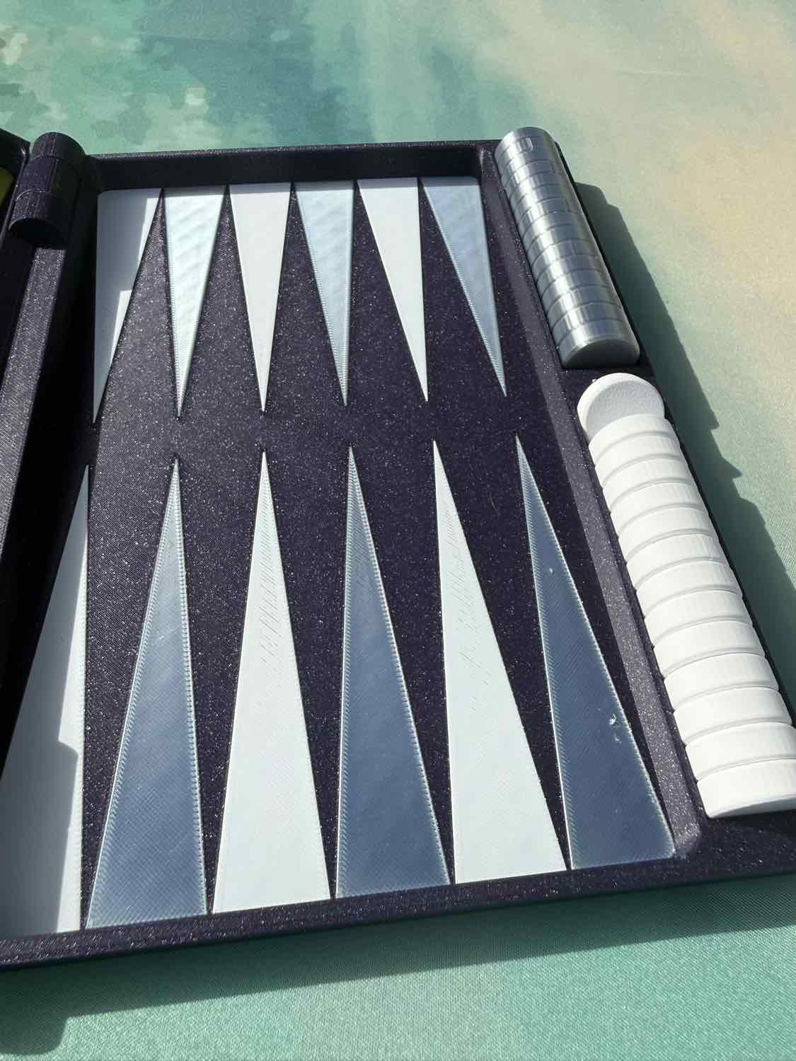 A close-up of a backgammon board with black and white triangular points, a black case, and white and silver checkers on a green surface.