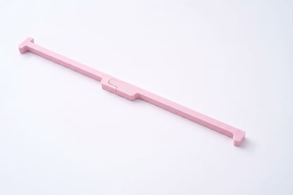 A light pink plastic tool with a long handle and a small central slider, photographed diagonally on a clean white background.