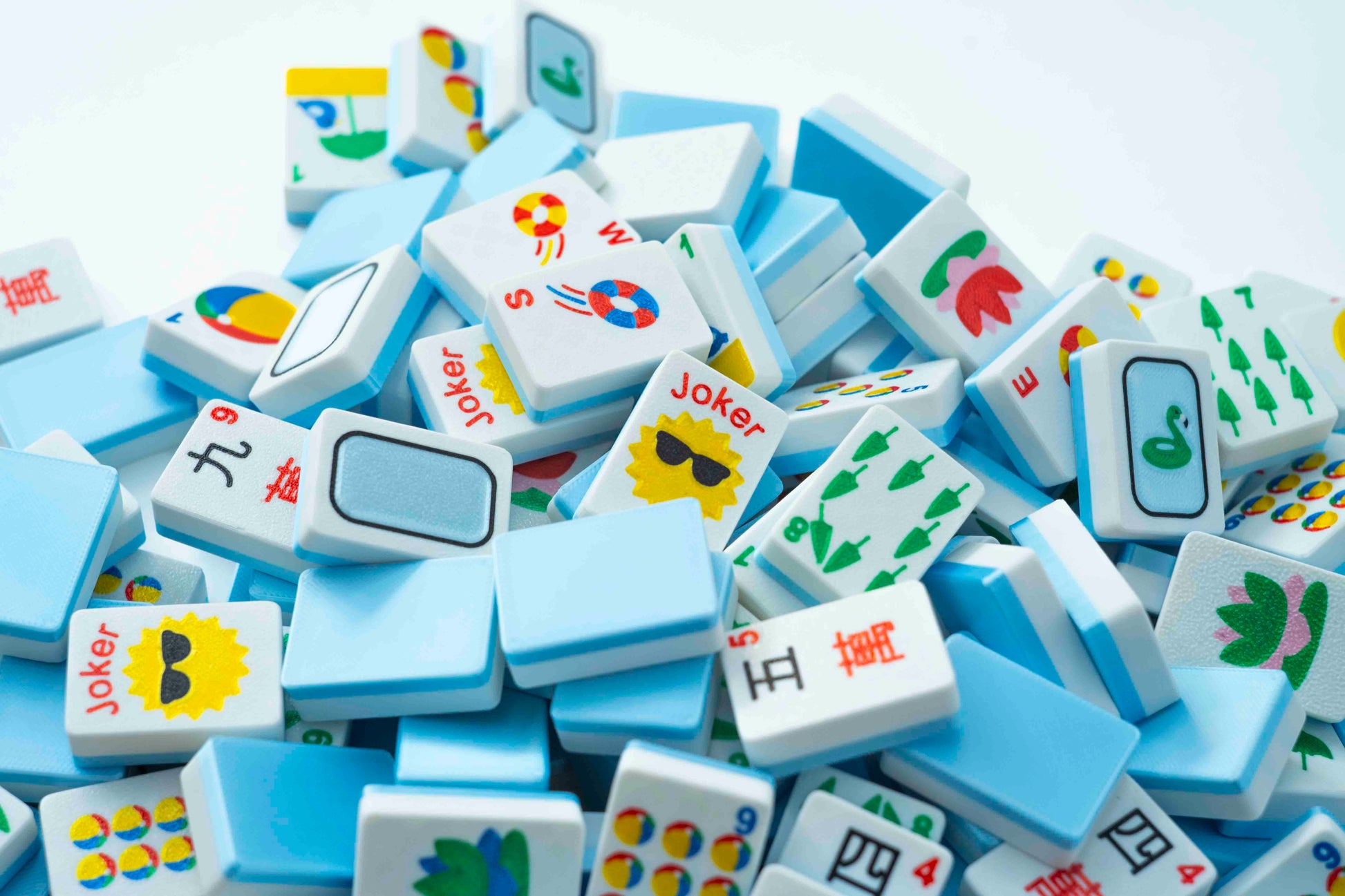 A pile of Mahjong tiles with white backs and colorful printed symbols, including Joker faces and Chinese characters, photographed on a light background.