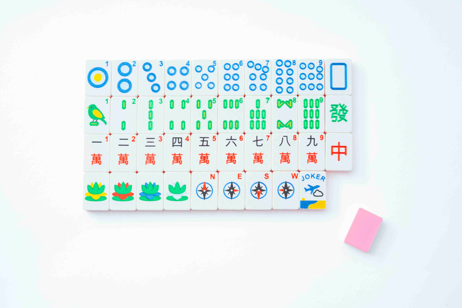 A set of Mahjong tiles arranged in rows on a white background, with a small pink eraser placed to the lower right. The tiles feature white bases with colorful Chinese gambling symbols in blue, green, red, yellow, and black.