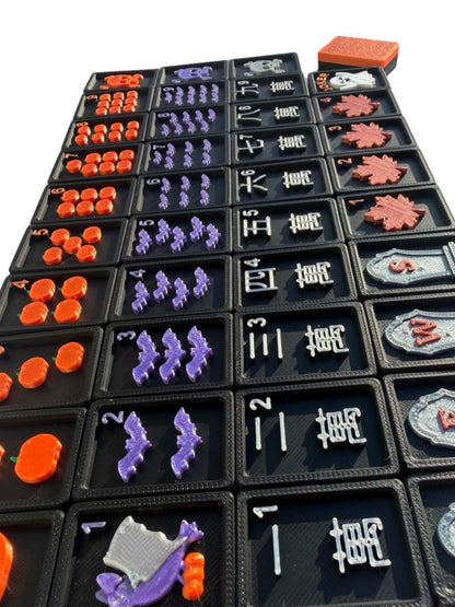 A close-up of a black and Orange Halloween Mahjong Tiless. The set includes orange, purple, white, and pink pieces arranged in columns, with some rectangular cards or tokens placed on top.