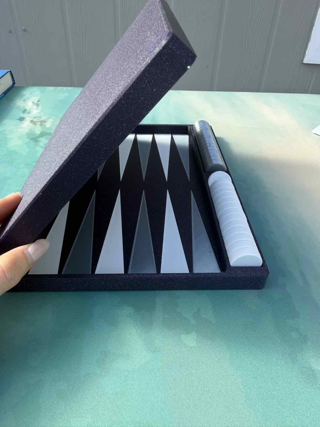 A hand holding open a dark gray foam presentation box containing a black and white backgammon board and a white-and-black cylindrical game piece, set on a mint green tabletop.