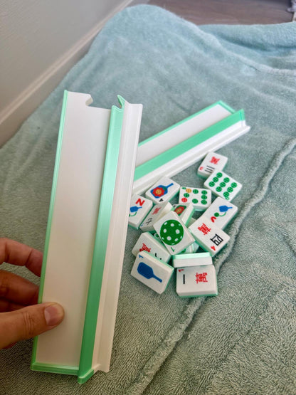 Hand holding mint green and white mahjong travel rack with floating pickleball-themed mahjong tiles scattered on a towel, including paddles, balls, and jokers