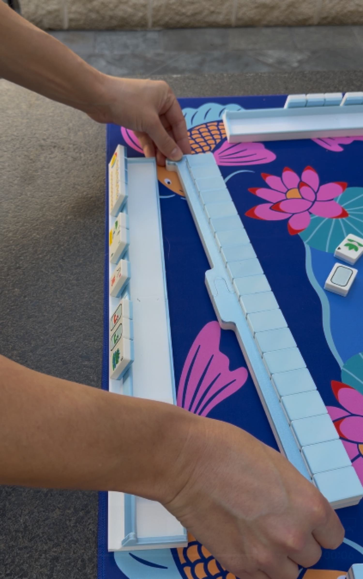 Hands assembling the blue Travel Mahjong rack and pusher on a lotus-pattern game mat, demonstrating the Spools travel-friendly setup
