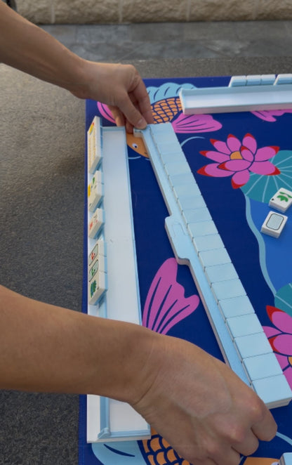 Hands assembling the blue Travel Mahjong rack and pusher on a lotus-pattern game mat, demonstrating the Spools travel-friendly setup