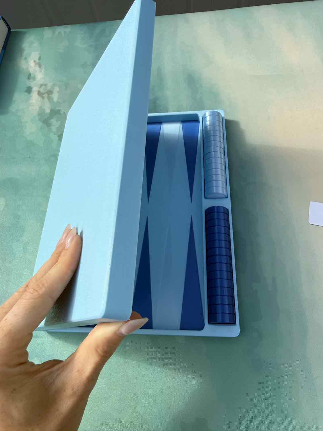 A hand holding open a light blue presentation box containing a blue and white backgammon set with a checkered die cup, placed on a green surface.