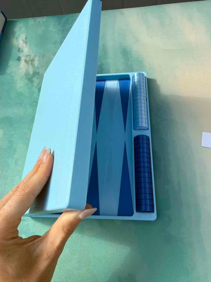 A hand holding open a light blue presentation box containing a blue and white backgammon set with a checkered die cup, placed on a green surface.