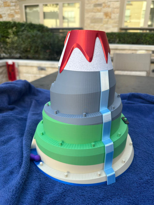 A 3D-printed volcano board game featuring layered tiers of green and gray slopes, a red lava peak, and a bright blue waterfall cascading down the mountain. Designed for racing pieces up the volcano in a fun, competitive tabletop game.